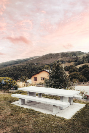 A vertical wide-angle shot of a stone picnic table with benches on grassy meadow overlooking countryside house, blooming flowers, forested hills and mountains under vibrant pink sunset skyの写真素材