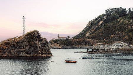 Coastal Cudillero harbor with moored fishing boats, rocky islet and lighthouse on a rugged headland, a busy pier and pastel sky reflecting on calm water - atmospheric maritime village scene; Asturiasの写真素材