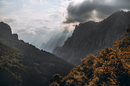 Dramatic sun rays pierce cloud over jagged peaks of Picos de Europa, revealing misty layered valleys and autumn-tinted forest on a rugged mountain landscapeの写真素材