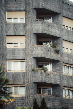 Modern residential building facade with dark brick walls, balconies decorated with plants, and large windows creating urban architectural detail in contemporary city housing; vertical telephoto shotの写真素材