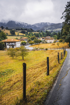 Picturesque rural village in Asturias during autumn with green and golden fields, traditional houses with tiled roofs, misty mountains in the background, and a wet country roadの写真素材