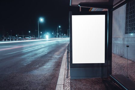 Blank illuminated advertising board at a bus shelter beside a wet city road at night, captured with long exposure car light trails and glowing street lampsの写真素材
