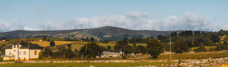 Wide autumn panorama of rural Asturias with a yellow country house in the foreground, rolling green and golden fields, scattered farms, and a distant ridge lined with wind turbines under a blue skyの写真素材