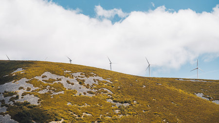 Row of wind turbines on a mountain ridge under a partly cloudy blue sky, blades turning above rocky slopes with golden vegetation. Clean energy and sustainable power in natural landscapeの写真素材