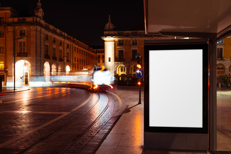 Blank vertical billboard mockup at a bus stop in Lisbon at night, illuminated by warm city lights and blurred tram motion on wet cobblestone tracks, perfect for urban advertising designの写真素材