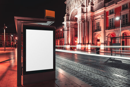 Night cityscape featuring a blank vertical billboard at a modern bus stop with street lights, motion blur of passing traffic, and illuminated historic architecture in downtown Lisbon, Portugalの写真素材