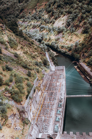Aerial view of a hydroelectric power station built into a steep green canyon, with transmission lines, concrete dam, and river flowing below, symbolizing renewable energy and engineeringの写真素材