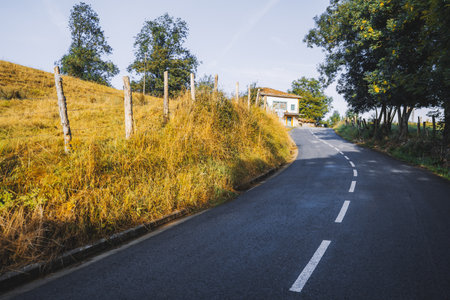 Curved rural road leading uphill through golden autumn grass and trees, with a small house at the top under clear blue sky, peaceful countryside landscapeの写真素材