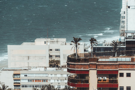 Modern beachfront apartment buildings with palm trees on terraces overlooking turquoise ocean waves, captured in bright daylight from elevated viewpoint in Arpoador Ipanema areaの写真素材