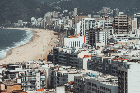 Telephoto view of Ipanema Beach with tall modern buildings, golden sand, and turquoise ocean waves stretching along the coast, framed by lush green mountains and vibrant city lifeの写真素材