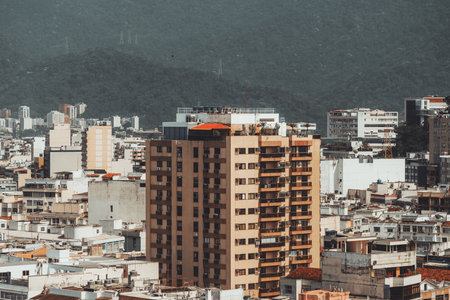 Urban cityscape with tall residential apartment building surrounded by dense city blocks, modern architecture, with green mountain forest in background under hazy daylight atmosphere; Rio de Janeiroの写真素材