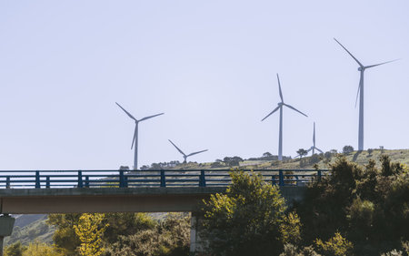 Row of modern wind turbines on green hillside behind highway bridge, clean renewable energy...