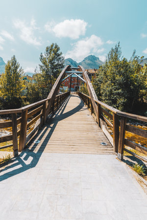 Vertical wide-angle view of a modern wooden pedestrian bridge in Cabrales, Asturias, leading toward mountains under blue sky, surrounded by green trees and warm sunlight, scenic travel destinationの写真素材