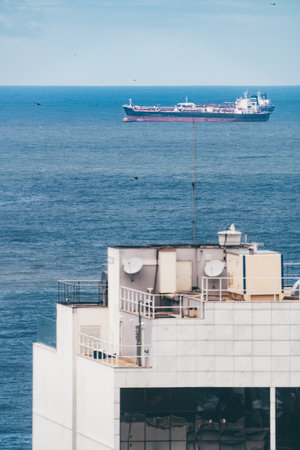 Cargo ship sailing on calm blue sea viewed through telephoto lens, with modern building rooftop in foreground featuring antennas, satellite dishes, and ocean horizon backgroundの写真素材
