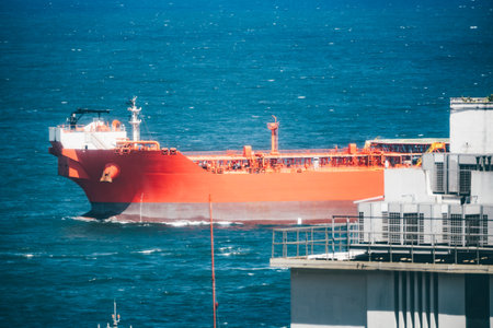 Large bright red oil tanker sailing in deep blue ocean water near an industrial port, captured with a telephoto lens showing ship details and modern building structures in foregroundの写真素材