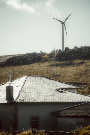 Selective focus telephoto view of rural landscape with house rooftops in foreground and large wind turbine on grassy hill in background, symbolizing renewable energy in countryside settingの写真素材