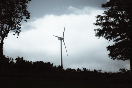 Silhouette of a modern wind turbine standing on a hilltop against dramatic cloudy sky, framed by dark trees and vegetation, symbolizing clean renewable energy and sustainabilityの写真素材