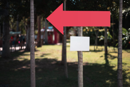 Blank mockup sign with large red arrow pointing left attached to a tree in sunny park, ready for custom text or logo placement, concept of outdoor direction and signage designの写真素材