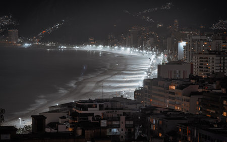 Telephoto long exposure of Ipanema beach at night, illuminated shoreline leading toward Arpoador and distant Leblon, blurred ocean wave motion and city light reflections creating a moody coastal skylineの写真素材