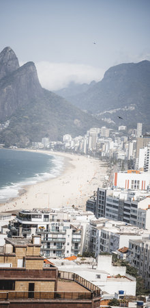 Vertical panorama of Ipanema beach with views toward Arpoador and distant Leblon, showing sandy shoreline, modern buildings, and dramatic mountains under soft daylight hazeの写真素材