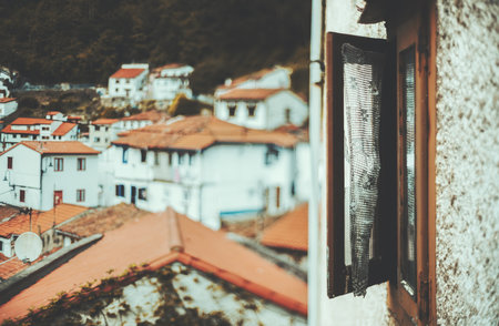 Rustic open window with torn curtain overlooking picturesque Cudillero village, Asturias, Spain, with charming white houses and red tiled roofs; selective focus with tilt-shift under natural daylightの写真素材