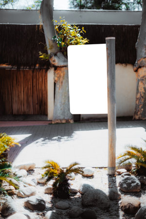 Outdoor mockup scene with blank white vertical signboard on wooden pole, surrounded by small palm plants, stones and soft sunlight, perfect for branding or advertising designの写真素材