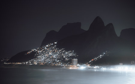 Long exposure telephoto night view of Vidigal and Leblon beneath Dois Irmaos peaks, Rio de Janeiro, with shimmering lights reflecting on the calm Atlantic Ocean under dark skyの写真素材