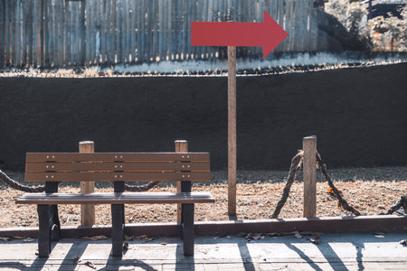 Blank red arrow sign mockup on wooden pole beside empty bench in sunny outdoor park, perfect template for direction, navigation or signage design presentation with natural backgroundの写真素材