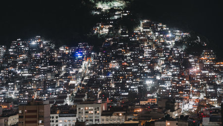 Telephoto view of illuminated favela community at night covering hillside with dense urban housing and glowing windows, symbolizing city life, population density and social contrastの写真素材