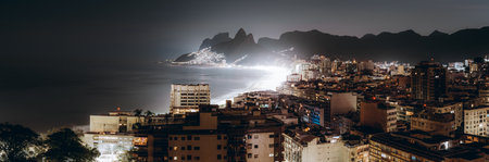 Panoramic night view of Arpoador, Ipanema and Leblon coastline with illuminated beach, ocean waves and mountain silhouettes, stunning Rio de Janeiro urban seascape under city lightsの写真素材