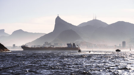 Large cargo ship and small boats sailing across Guanabara Bay with Rio de Janeiro skyline and iconic mountain silhouette under hazy afternoon light and sparkling sea reflectionsの写真素材