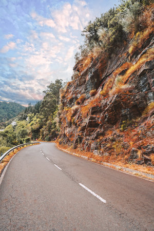 Wide-angle shot of a curved asphalt road winding through colorful rocky mountainside in Asturias, Spain, along the scenic Camino de Santiago under dramatic cloudy sky during autumn season travelの写真素材