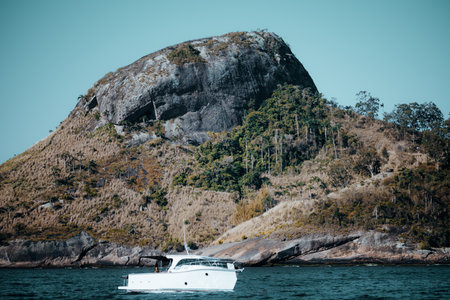 White motor yacht cruising near rocky tropical island off Rio de Janeiro, Brazil, with steep green hillside and clear turquoise water under bright summer sunlightの写真素材