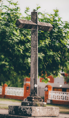 Ancient stone cross standing on a weathered pedestal, captured in vertical telephoto composition. Soft light highlights moss and lichen texture with blurred trees and village backgroundの写真素材