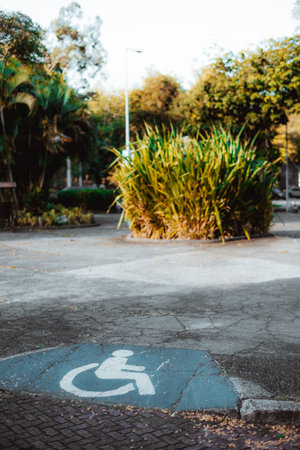 Vertical shot of a wheelchair accessibility ramp with blue disabled parking symbol on cracked pavement, shallow depth of field showing greenery and soft sunlight in the urban park backgroundの写真素材