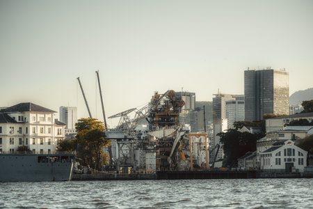 Huge offshore construction vessel with towering cranes and complex machinery docked at urban harbor for maintenance; framed by old white warehouses, modern skyscraper skyline and rippling waterの写真素材