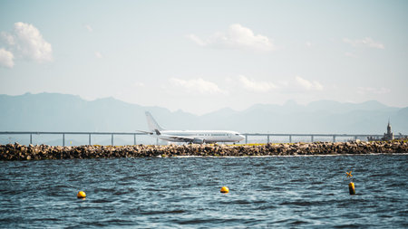 Commercial airplane taxiing along a rocky shoreline with distant mountains, calm sea water and bright marker buoys under clear sky in a long-lens telephoto coastal sceneの写真素材