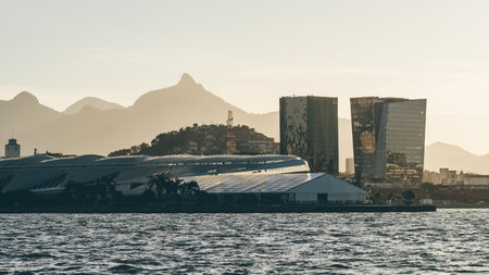 Modern waterfront skyline with reflective high-rise towers, futuristic roof structures and distant mountain silhouettes glowing in warm sunset haze above rippling coastal waterの写真素材