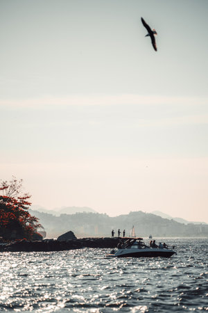 Silhouettes of distant unrecognizable people on a rocky shoreline and a boat gliding over shimmering...