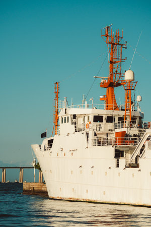 Brightly lit white research vessel with vivid orange masts docked by calm waterfront, showcasing detailed maritime equipment and clean geometric lines against a clear blue skyの写真素材