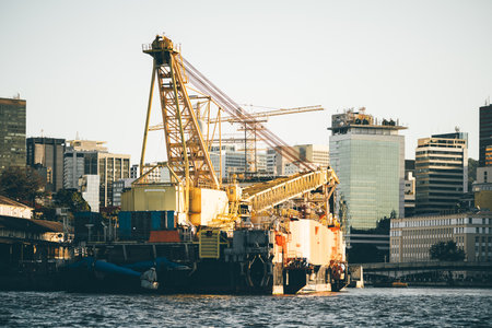 Large industrial floating crane platform with towering yellow structures, heavy equipment and cables; against a dense skyline of modern office buildings at golden hour on calm waterfront; Rio, Brazilの写真素材