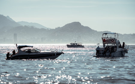 Telephoto scene of leisure boats on shimmering water with distant hazy coastal skyline and layered mountains, capturing relaxed seaside atmosphere and sparkling midday light and silhouettes of peopleの写真素材