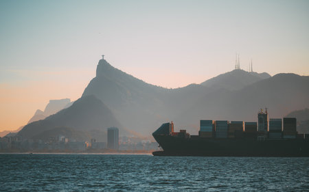 Majestic telephoto coastal view with silhouetted cargo ship passing before Rio mountains at soft sunrise hues, revealing city skyline layers and the iconic peak crowned by a towering statueの写真素材