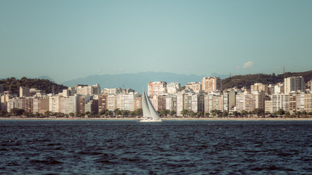 Telephoto view of Niteroi coastline with dense beachfront skyline, calm sea and a white sailboat gliding in foreground against distant soft-blue mountain silhouettesの写真素材