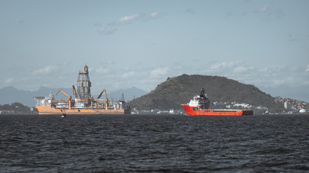 Offshore drilling rig and red supply vessel on a coastal bay, industrial oil and gas operation with cranes and derrick, distant shoreline city and mountain under wide blue skyの写真素材