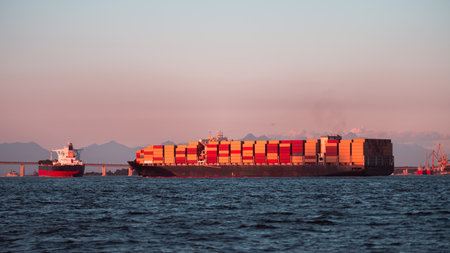 Large container ship loaded with colorful containers sailing at sunset on calm sea, with tanker, bridge and port cranes in distance, global maritime freight transportの写真素材