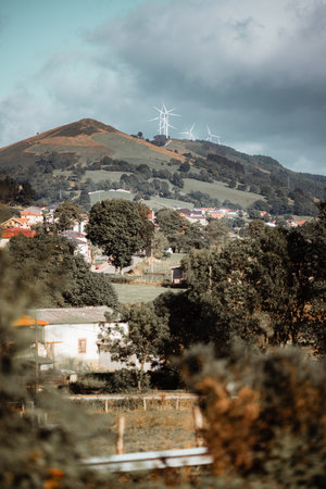 Telephoto layered countryside view: blurred foliage foreground, village and field midground, green...