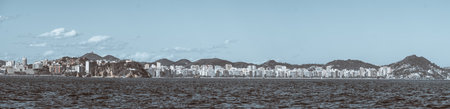 Telephoto panoramic view of Niteroi waterfront skyline across Guanabara Bay, modern apartment towers and green hills under blue sky, textured sea foreground, copy spaceの写真素材