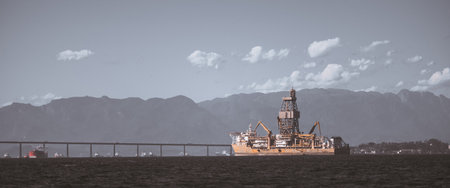 Telephoto view of offshore oil drilling platform ship near a long bridge with hazy mountain backdrop, calm sea, industrial energy concept, wide copy space on the leftの写真素材