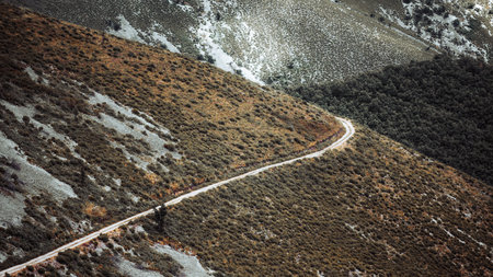 Winding dirt road crossing rugged hillside with scrub vegetation and rocky patches, aerial landscape view of remote mountain terrain, natural texture background and copy spaceの写真素材